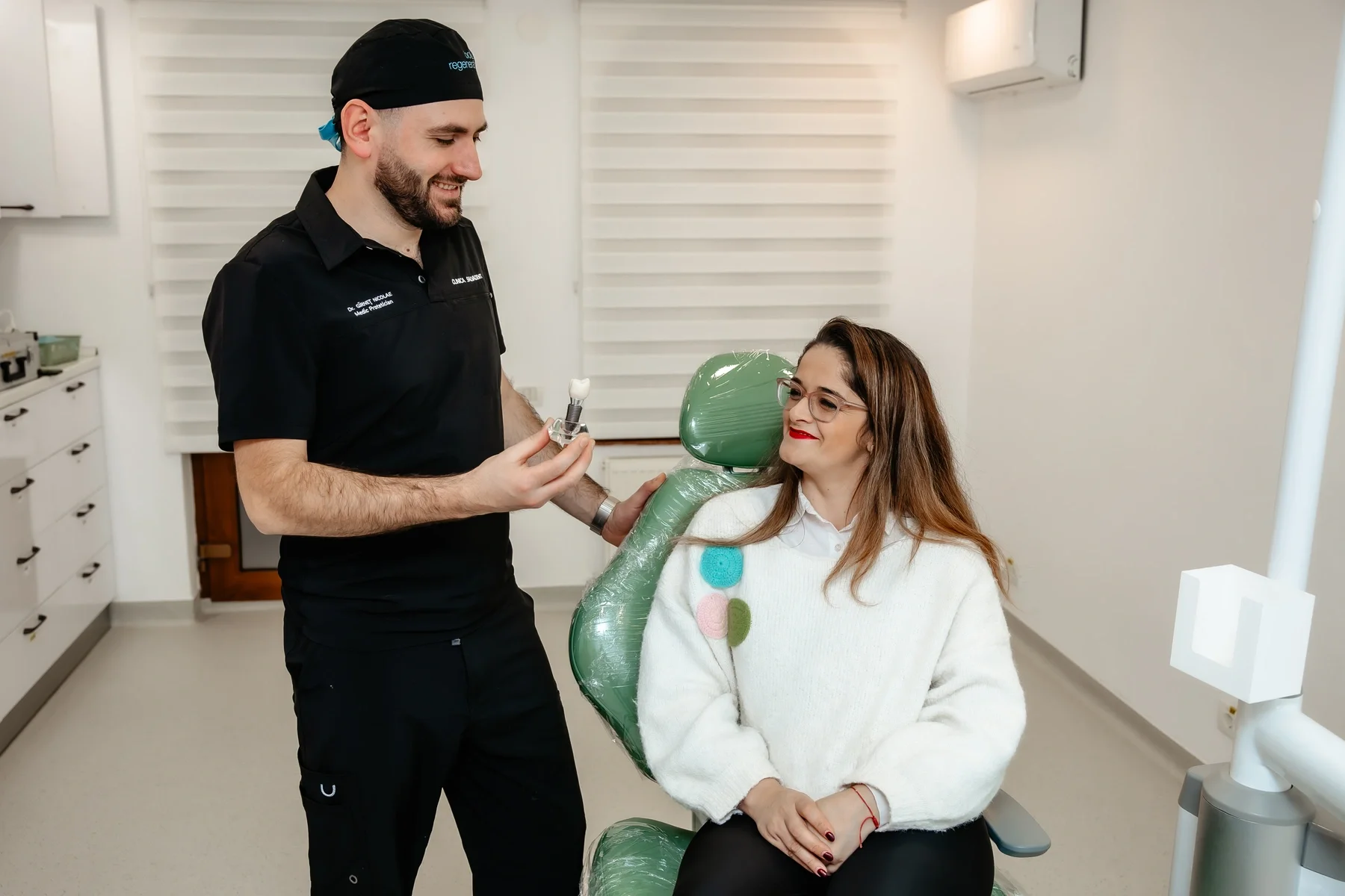 Medic stomatolog arătând unei paciente un model de implant dentar, în cabinet