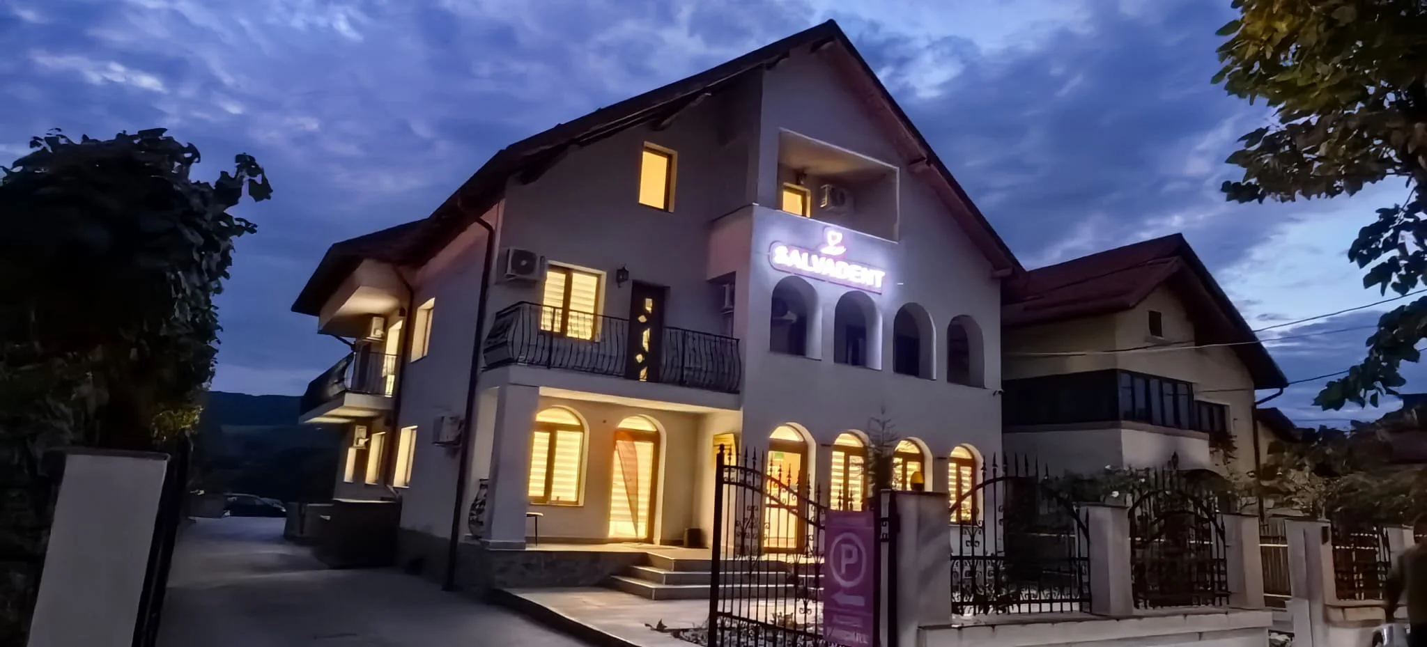 Exteriorul Clinicii SalvaDent din Curtea de Argeș la apus, cu firma luminoasă deasupra intrării